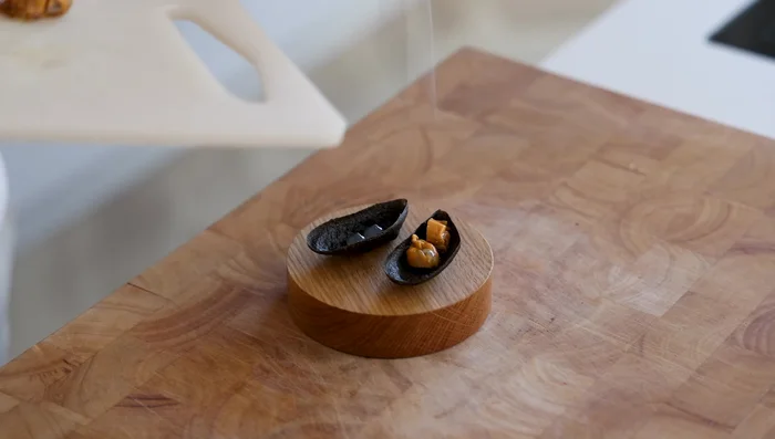 To assemble, pipe two small dots of gel onto the bottom of each pie tee, top with a portioned mussel, flaky salt, coconut foam, and garnish with green leaves.