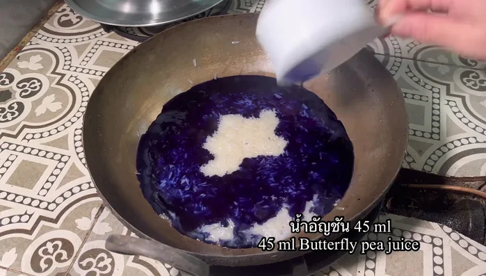After 15 minutes, heat the pan again. Add concentrated butterfly pea flower water. Gently stir in the rice, ensuring the color permeates evenly. Continue stirring in the same direction until the rice starts to clump and come off the pan.
